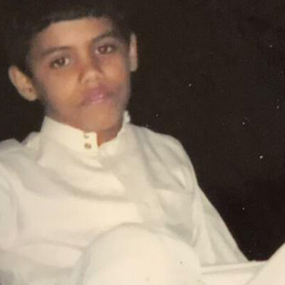 An image of the top half of a young Abdullah al-Howaiti looking directy at the camera. He is wearing a white Thawb which is an ankle length robe. He is sat against a dark black background. 